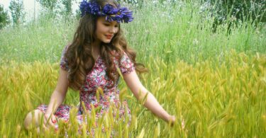 Imagem de uma mulher jovem sentada em meio a um campo e em sua cabeça uma coroa de flores roxas.