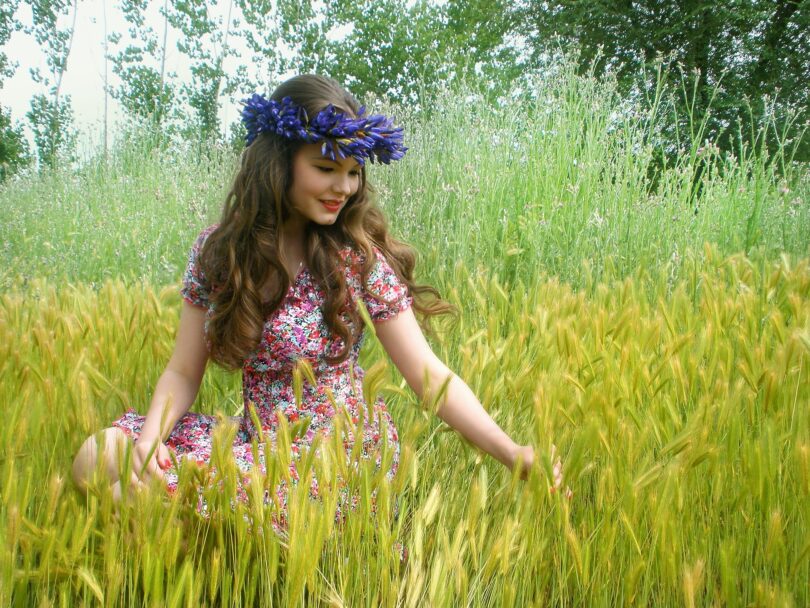 Imagem de uma mulher jovem sentada em meio a um campo e em sua cabeça uma coroa de flores roxas.