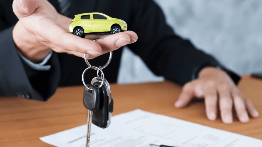 Homem segurando um carro em miniatura na mão, juntamente com a chave do veículo de verdade. Ao fundo, vemos um contrato de venda.