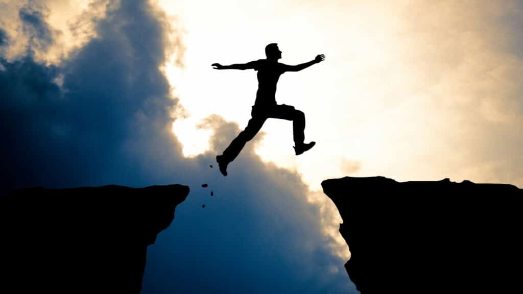 Imagem de um homem pulando de uma rocha para a outra e o céu azulado como paisagem