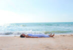 Mulher deitada no chão de areia em frente ao mar com o corpo para cima.