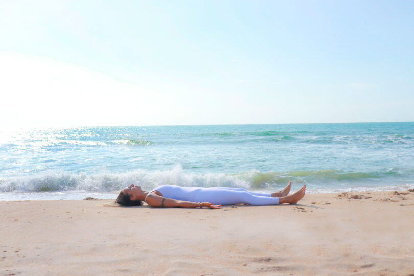 Mulher deitada no chão de areia em frente ao mar com o corpo para cima.