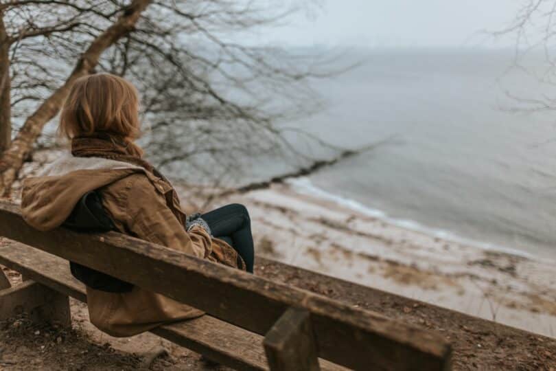 Mulher sentada em um banco, olhando o mar, em um dia nublado e frio.