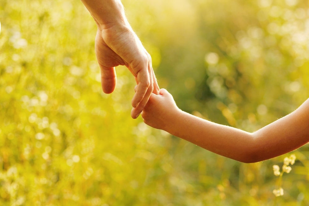 Mão de uma criança segurando a mão de um adulto em uma paisagem esverdeada.