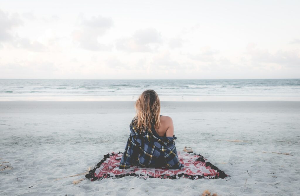 Mulher de costas, sentada na areia da praia, olhando para o mar.