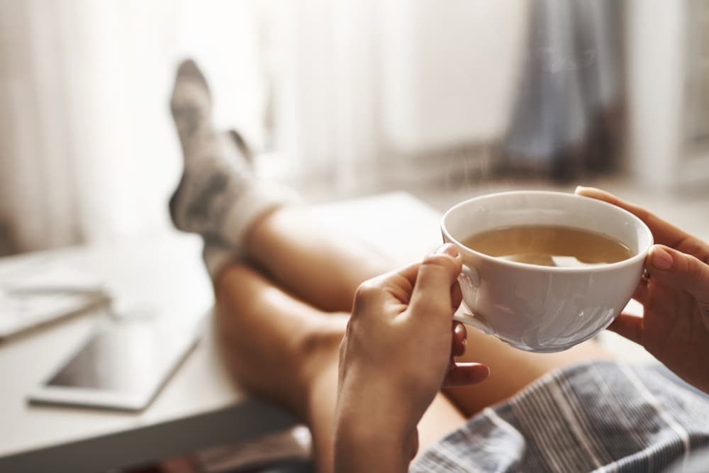 Mulher sentada, com as pernas estiradas e cruzadas, segurando uma xícara de chá.