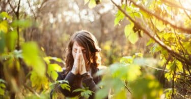 Mulher com as palmas das mãos unidas na frente de seu rosto, meditando com os olhos fechados. Ela está em meio à natureza.