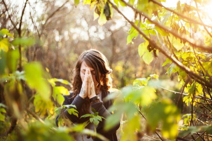 Mulher com as palmas das mãos unidas na frente de seu rosto, meditando com os olhos fechados. Ela está em meio à natureza.