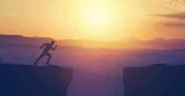 Imagem de um rapaz prestes a pular de uma rocha para a outra e uma paisagem de pôr do sol.