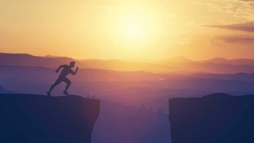 Imagem de um rapaz prestes a pular de uma rocha para a outra e uma paisagem de pôr do sol.