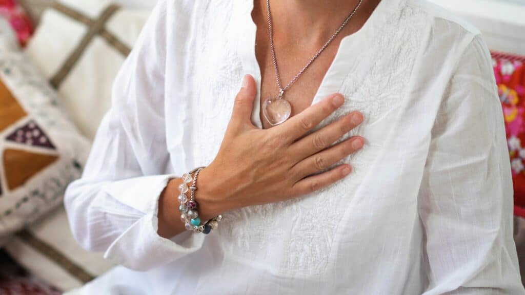 Imagem de uma mulher de branco, usando colar e pulseiras com a mão no coração.