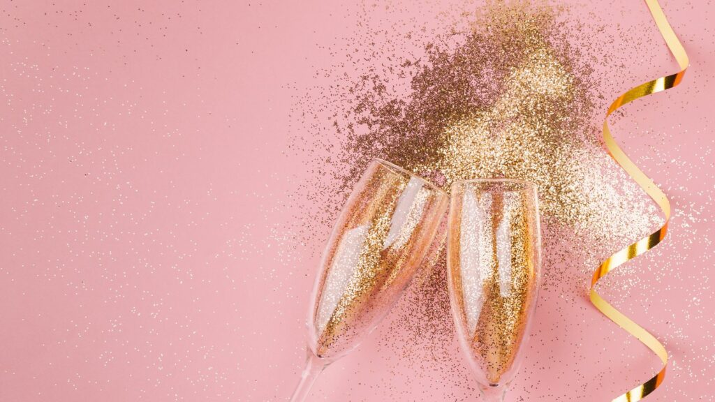 Imagem de duas taças com gliter dourado uma fita dourada de festa em um fundo cor de rosa