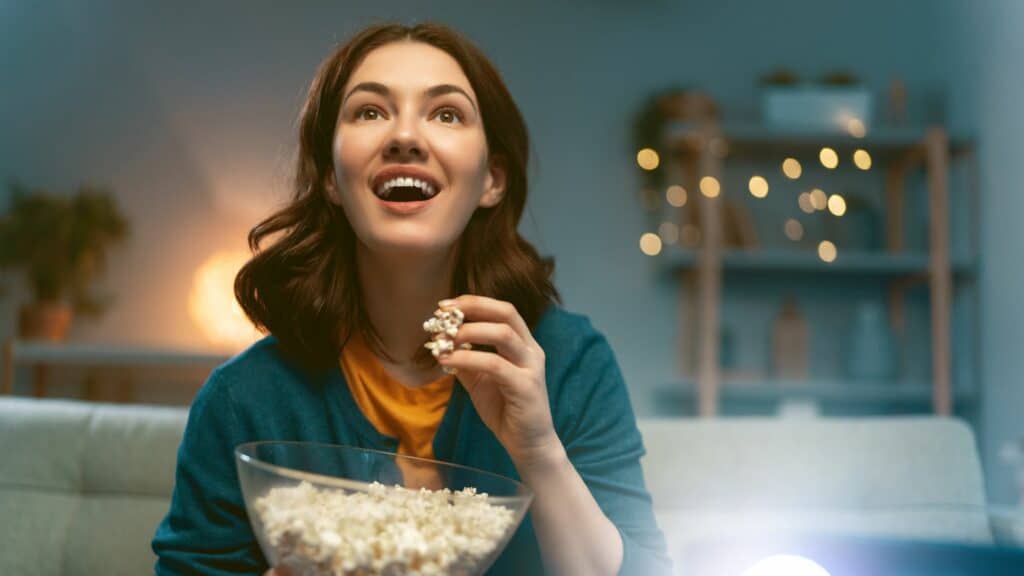 Imagem de uma mulher comendo pipoca e assitindo TV com a expressão de satisfação pelo o que vê.