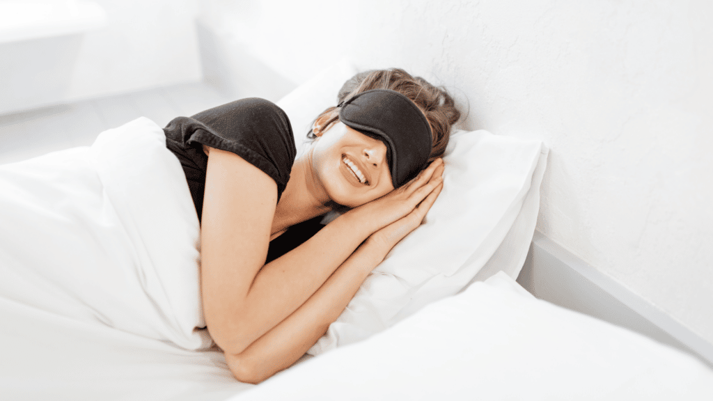 Mulher branca e de cabelos claros deitada na cama, usando tapa-olhos e sorrindo.