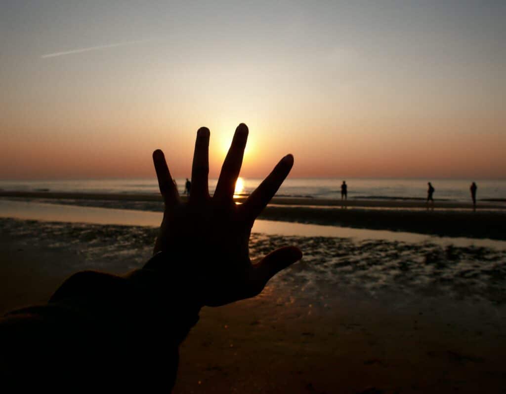 Mão sombreada, espalmada em direção ao mar. O Sol se põe no horizonte e há pessoas à beira do mar, ao longe.