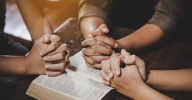Mãos de três pessoas sobre uma Bíblia, em posição de oração coletiva