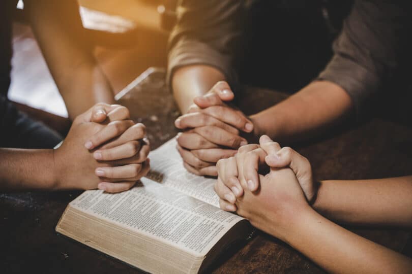 Mãos de três pessoas sobre uma Bíblia, em posição de oração coletiva
