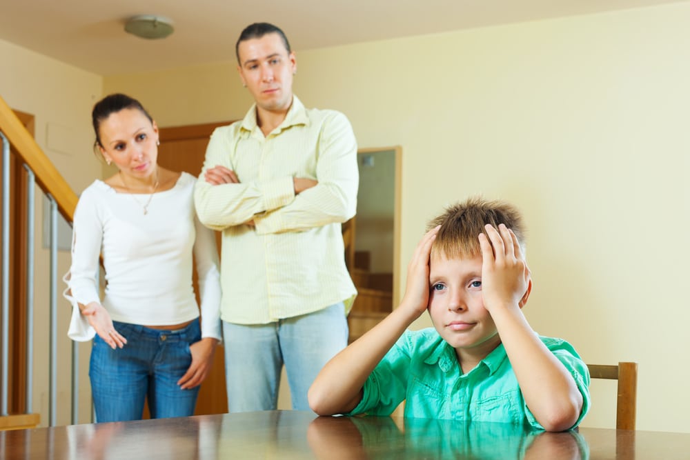 Casal de pais em pé, observando o filho sentado em uma mesa, com semblante frustrado.