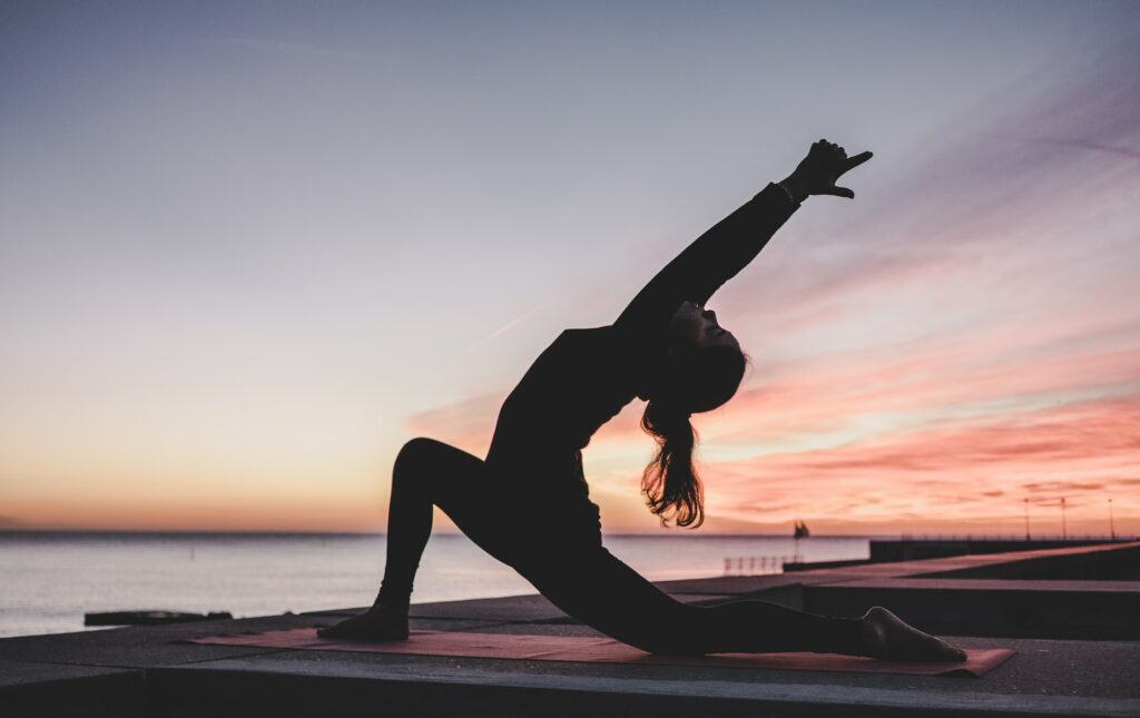 Mulher praticando uma postura de yoga à beira do mar, durante o pôr do sol.