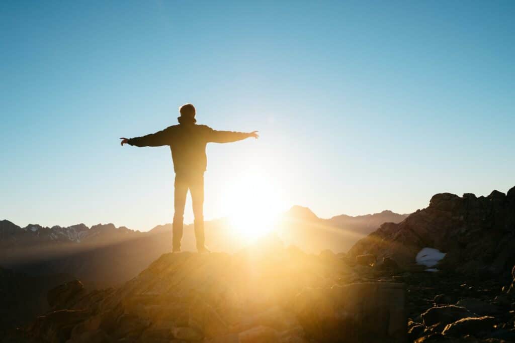 Homem em pé sobre uma superfície montanhosa, de braços abertos, de frente para o Sol.