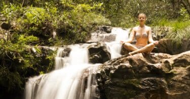 Imagem de uma mulher sentada perto de uma cachoeira fazendo Yoga