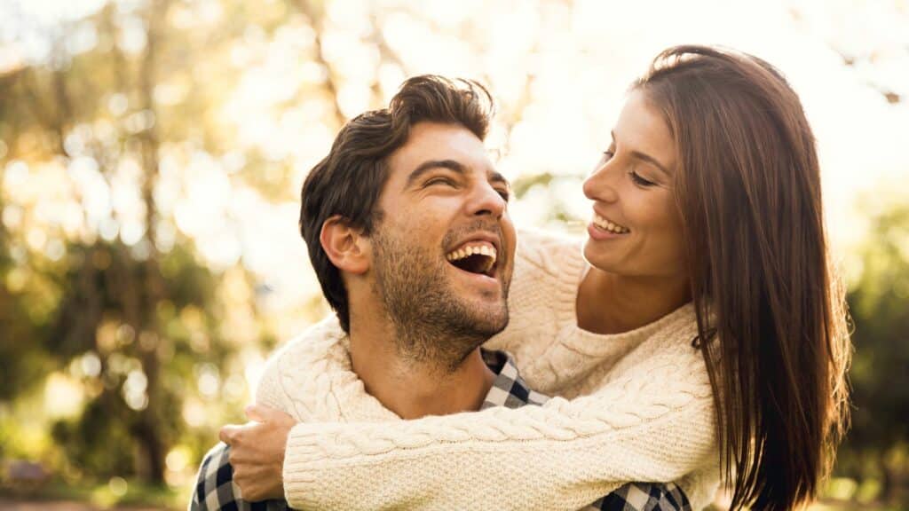Imagem de uma casal sorrindo, a mulher está abraçando o homem pelas costas