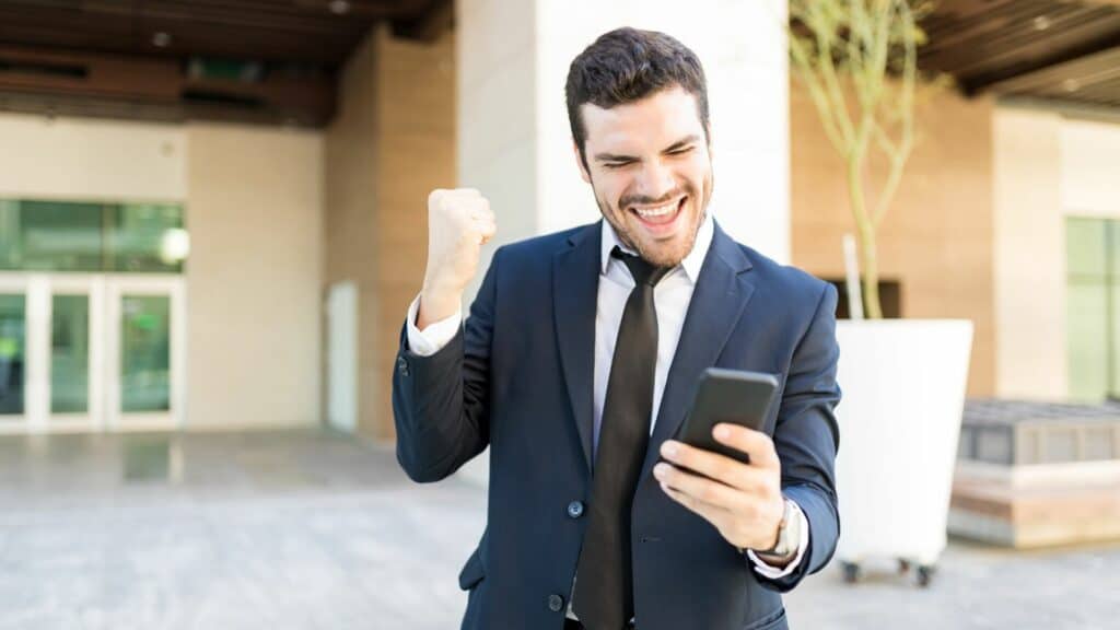 Imagem de um homem comemorando ao olhar no celular, como se tivesse recebido uma ótima notícia