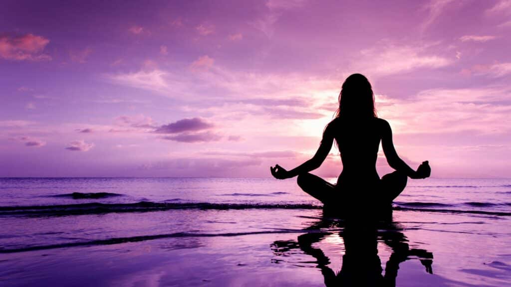 Imagem de uma mulher meditando na praia e o céu lilás como paisagem