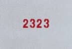 Número 2323 escrito na cor vermelha em uma parede branco acinzentada.