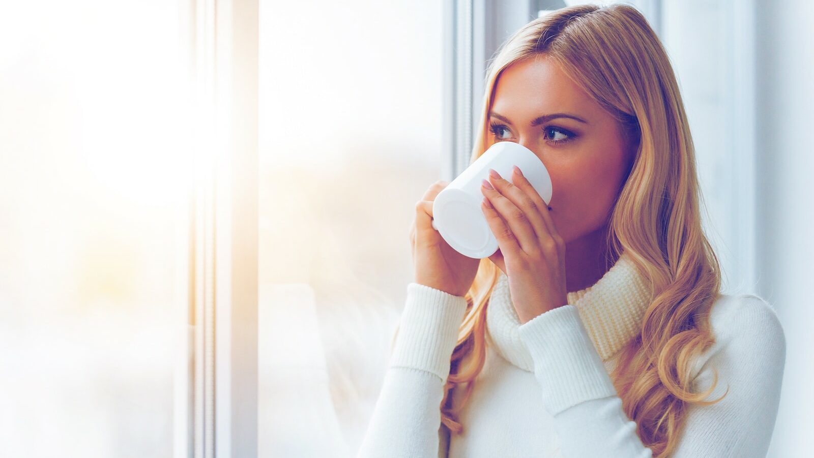 Enjoying fresh coffee. Beautiful young woman in white sweater drinking coffee and looking through a window