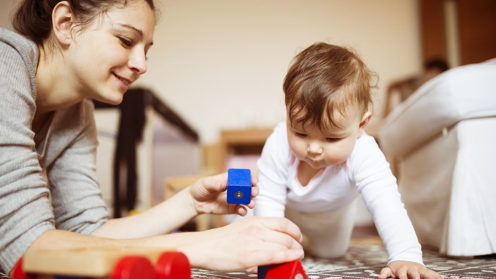 Cute little baby girl playing with her mother on a carpet in a living room.