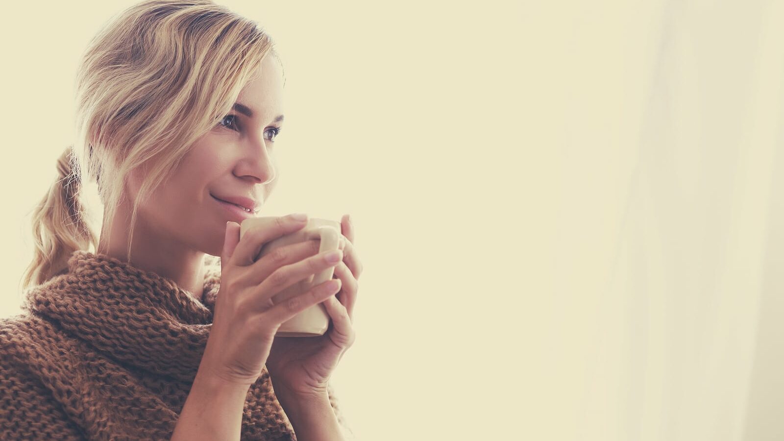 Woman wearing warm knitted sweater is drinking cup of hot tea or coffee near window in autumn morning sunlight, photo warm toned