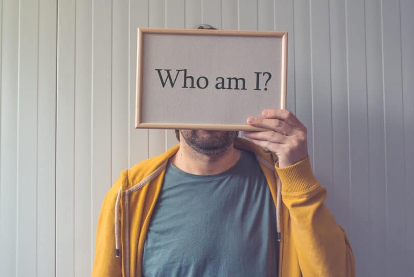 Homem segurando uma placa que cobre o seu rosto. Na placa, a pergunta: "who am I?"