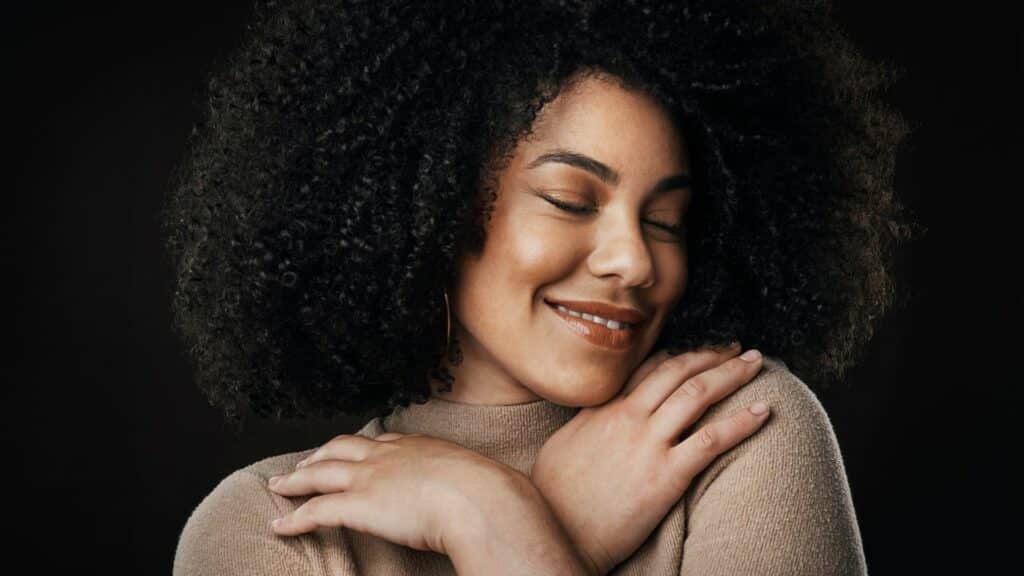Imagem de uma mulher sorrindo de olhos fechados e abraçando a si mesma em um fundo preto