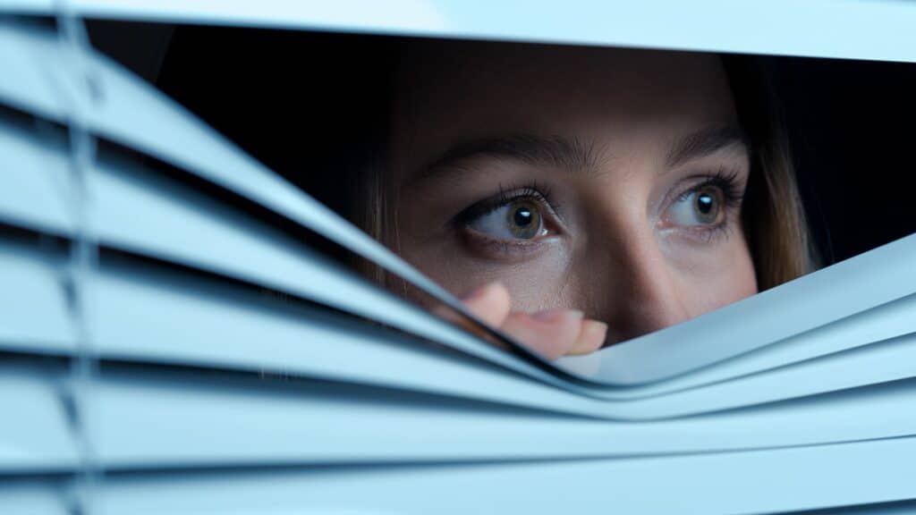Imagem de uma mulher olhando pela janela com medo como se não quisesse ser vista
