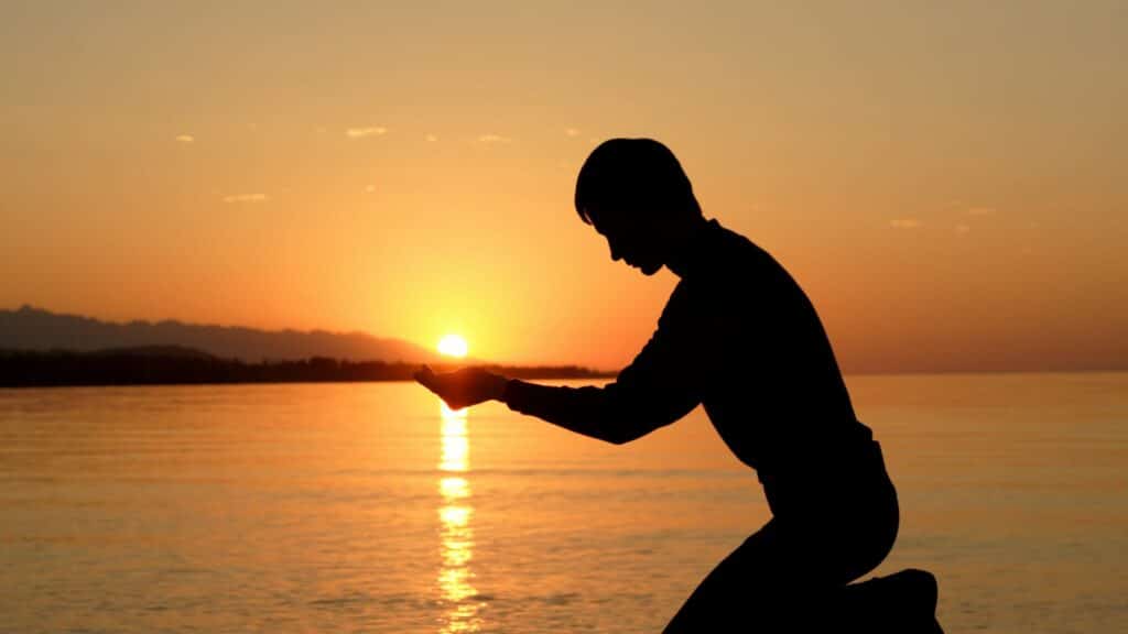 Imagem de um homem ajoelhado e atrás dele uma paisagem de pôr do sol e mar e suas mãos parecem segurar o sol.