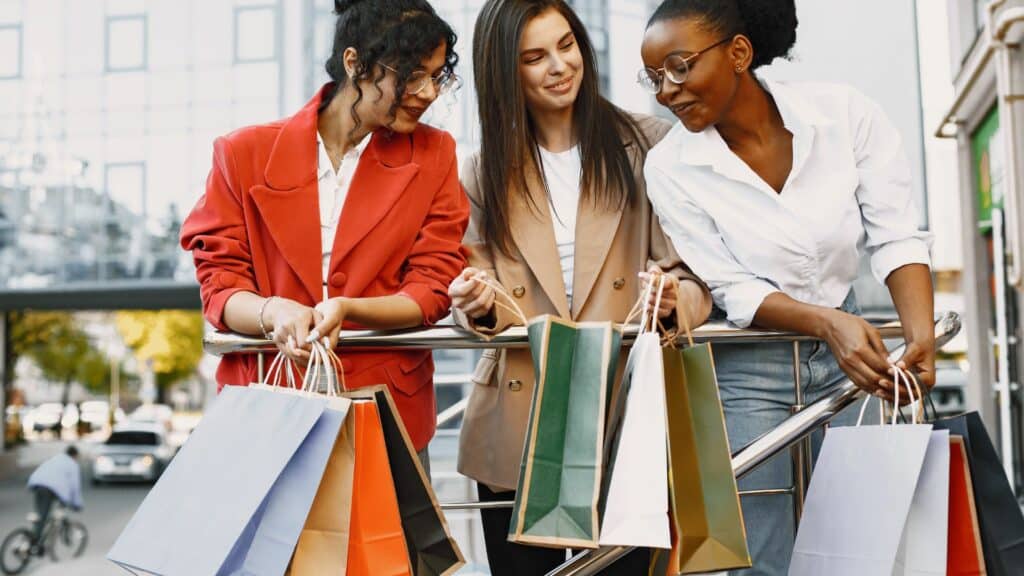 Imagem de três mulheres sorrindo, cada uma com sacolas de compras nas mãos