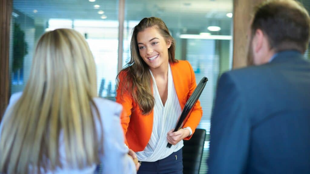 Imagem de uma mulher com trajes de trabalho sorrindo e se apresentando para um casal que parecem ser os chefes de uma empresa