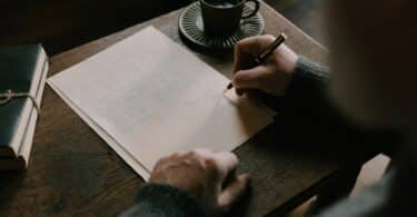 Imagem de um homem escrevendo em uma folha e ao seu lado, na escrivaninha, uma xícara de café
