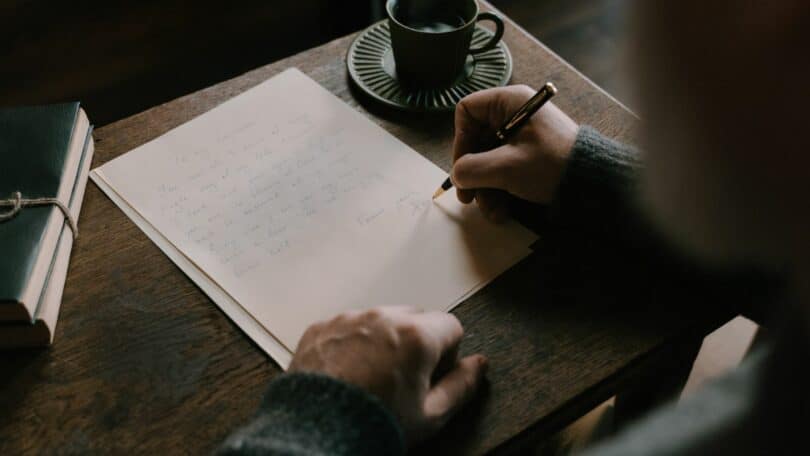 Imagem de um homem escrevendo em uma folha e ao seu lado, na escrivaninha, uma xícara de café