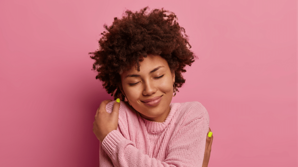 Mulher negra vestindo um suéter rosa de olhos fechados se abraçando