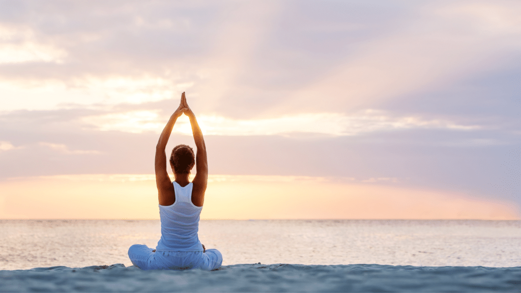 Mulher branca praticando yoga na praia, com o céu rosado ao fundo