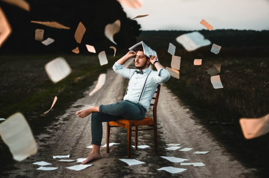 Homem sentado em uma cadeira, no meio de um caminho ao ar livre. Ele está descalço, segurando um livro aberto sobre a cabeça, e várias páginas soltas flutuam ao seu redor.