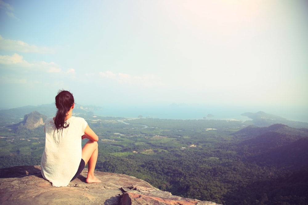 Mulher refletindo, sentada na beira de uma montanha.