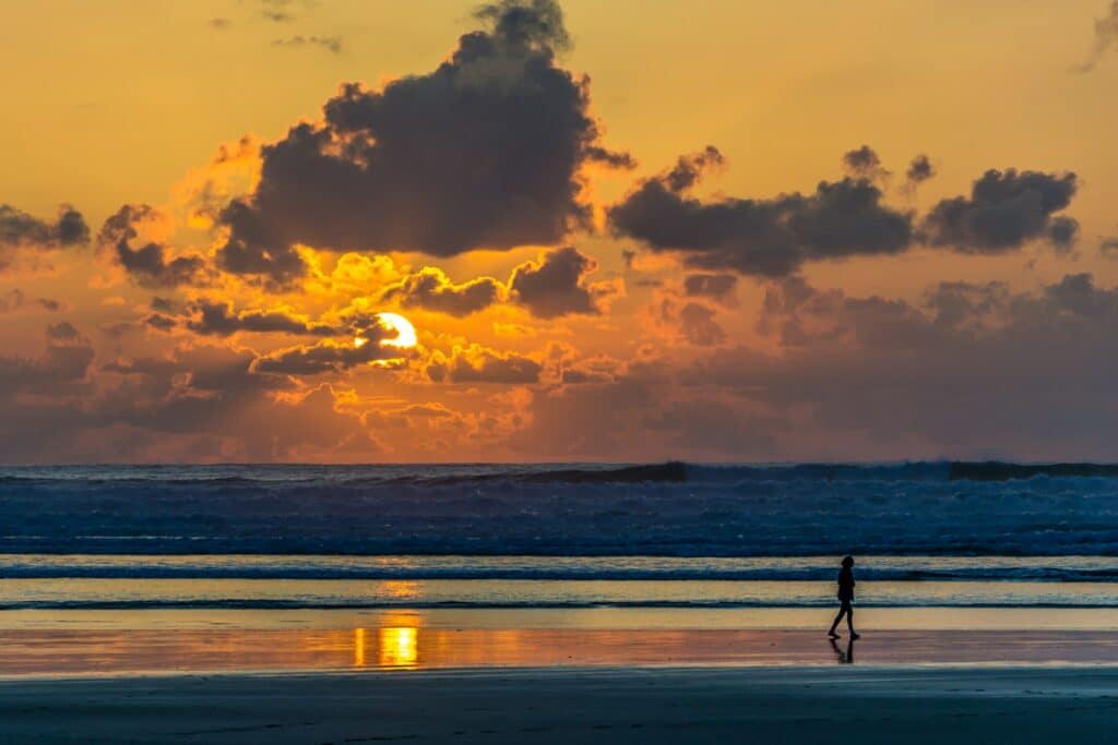 Silhueta de uma pessoa caminhando ao longe, na orla da praia, durante o pôr-do-Sol.