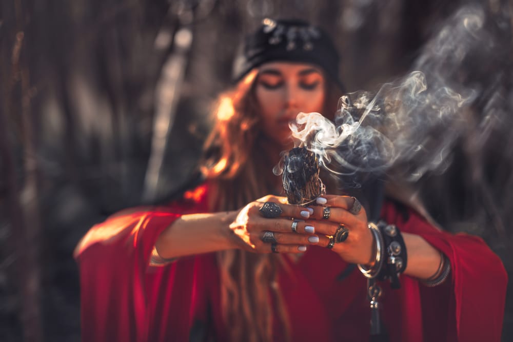 Mulher cheia de acessórios místicos em meio à floresta queimando um incenso