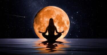 Silhueta de mulher sentada meditando sobre as águas com uma grande Lua dourada ao seu redor