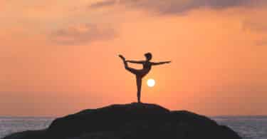 Sombra de uma mulher realizando yoga no topo de uma pedreira. O sol está se pondo no fundo