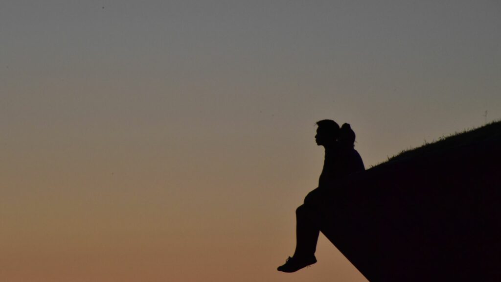 Imagem de um homem sentado na ponta de uma montanha, no final da tarde refletindo