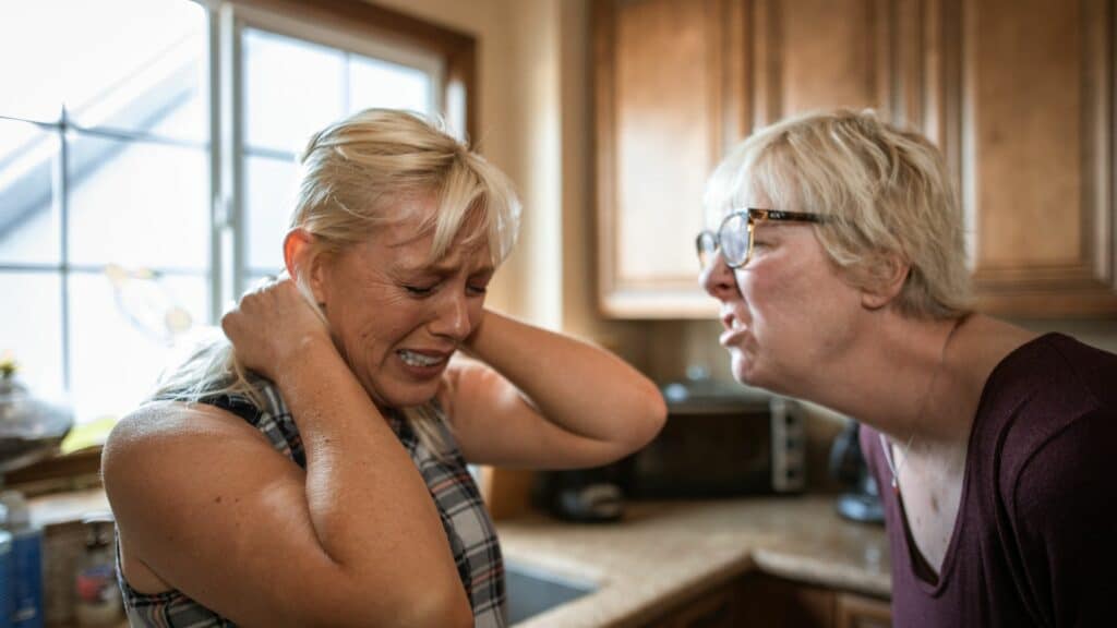 Imagem de uma mulher gritando e oprimindo outra em uma cozinha, a pessoa oprimida está chorando.